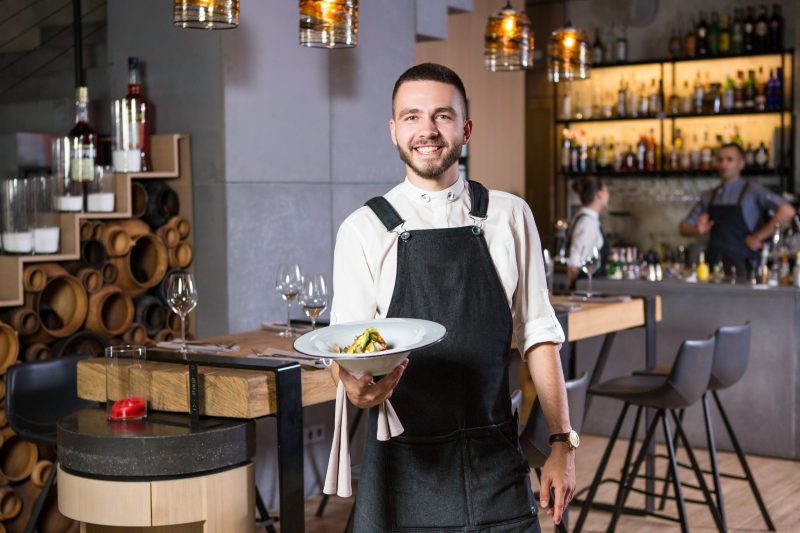 Male restaurant server standing in a restaurant holding a plate of food. The background is a high-top wood table with higher chairs with two servers behind the bar in the further back.